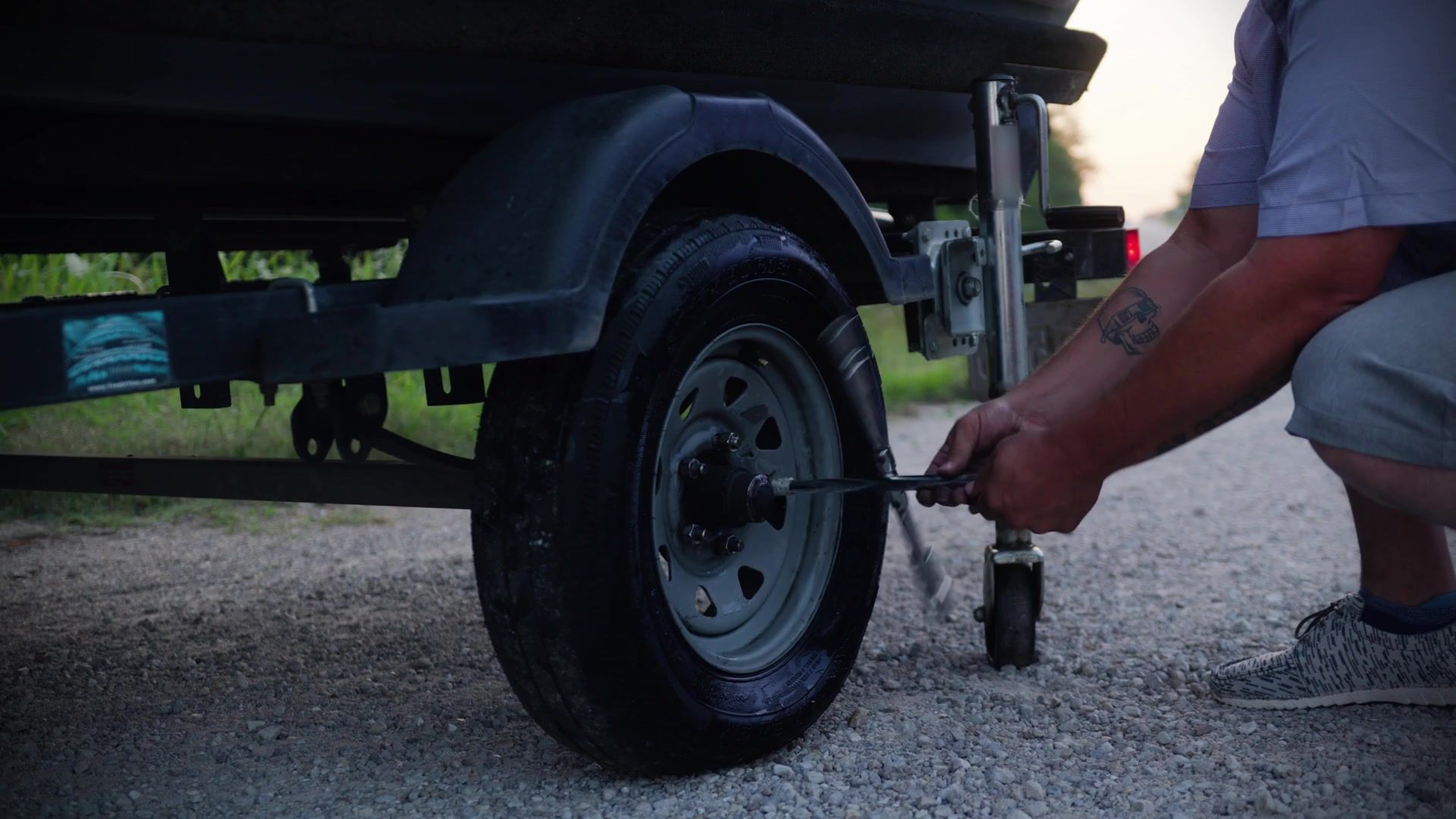 A man loosening the lug nuts on a trailer wheel lifted clear of the gravel, with the jack held in the Jack Bracket beside it